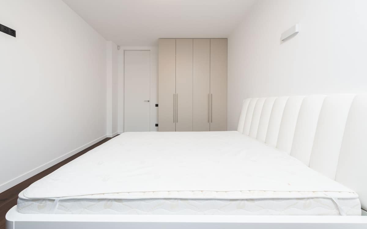 A bare mattress on a white bed frame with a quilted top layer laid over it